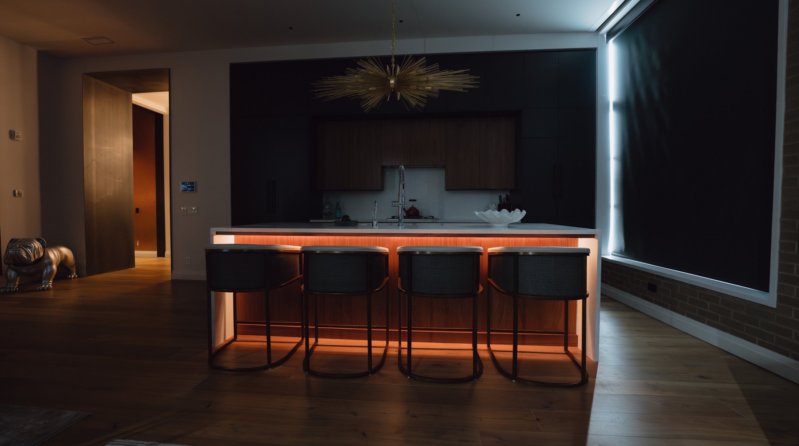 A residence kitchen at night with an amber-lit island, a motorized blackout shade lowered over the window, and a sculptural chandelier — by Partner Technology